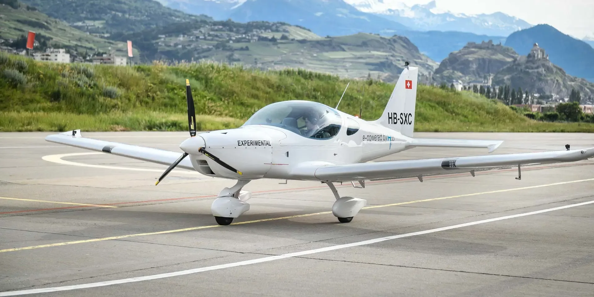 A white Pipistrel Velis Electro experimental aircraft, registered HB-SXC, sits on an airfield tarmac with green hills and mountains in the background.