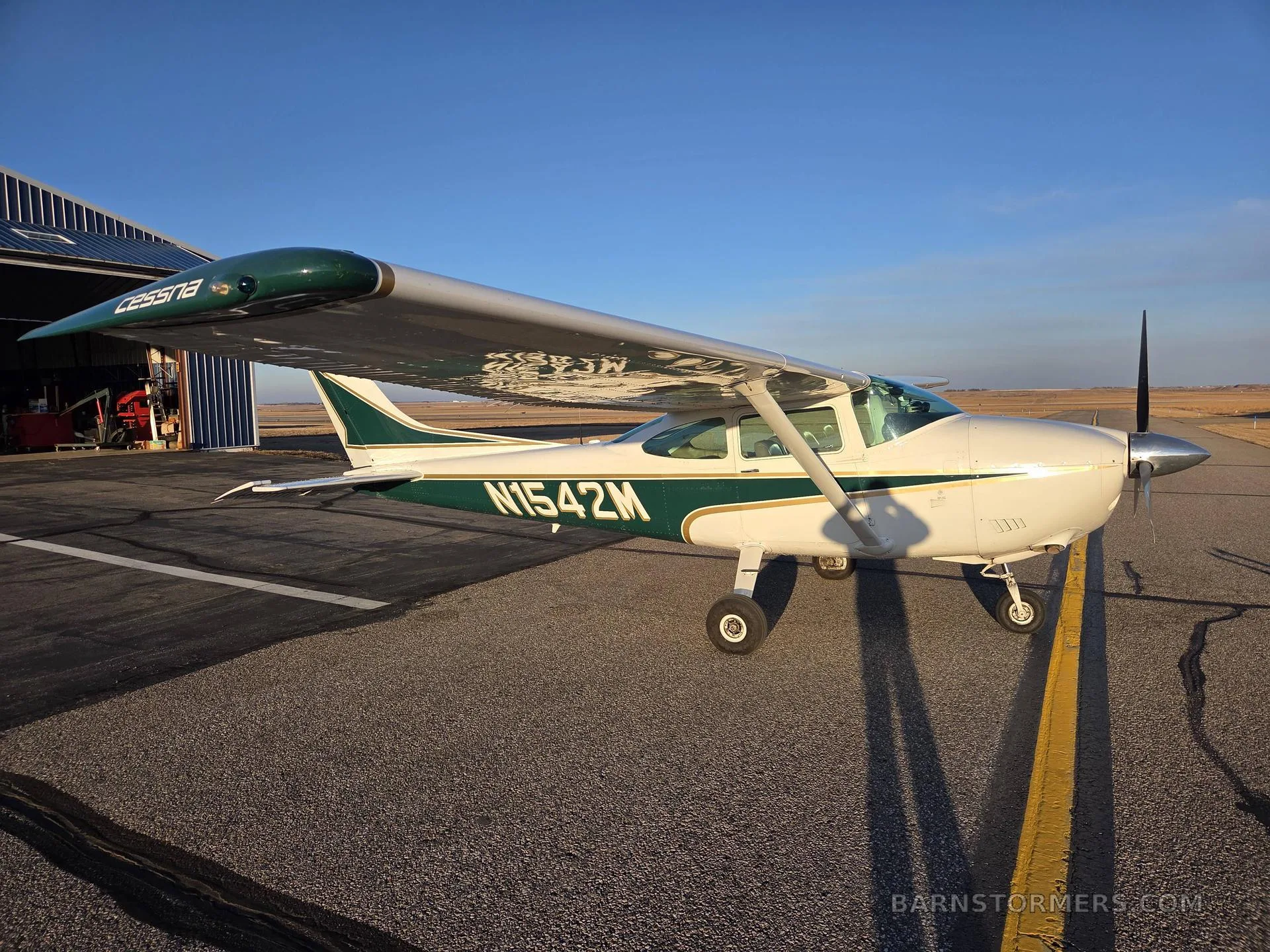 A white and green Cessna aircraft with registration N1542M parked on an airfield next to a hangar.