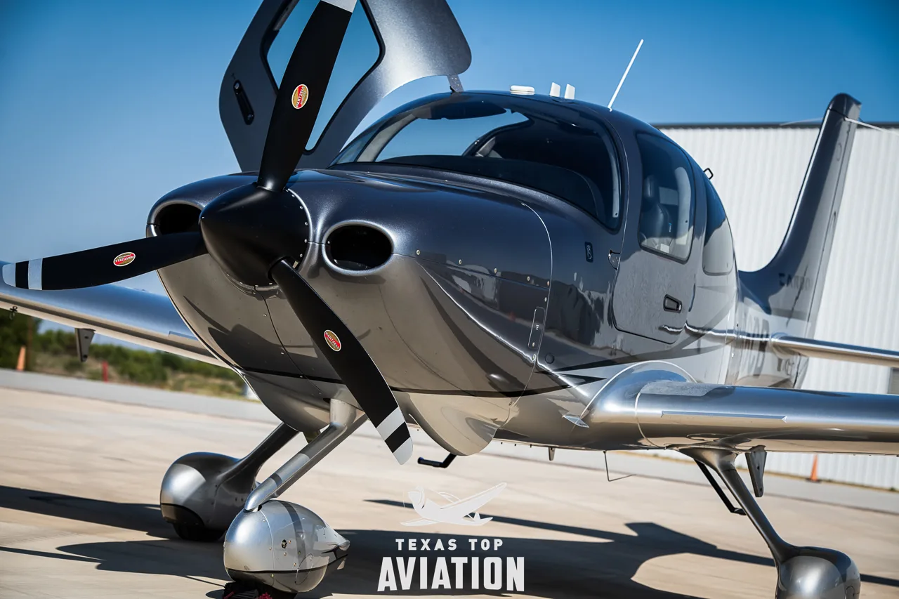 A close-up front view of a silver and grey Cirrus SR22 aircraft with its propeller and open cowl.