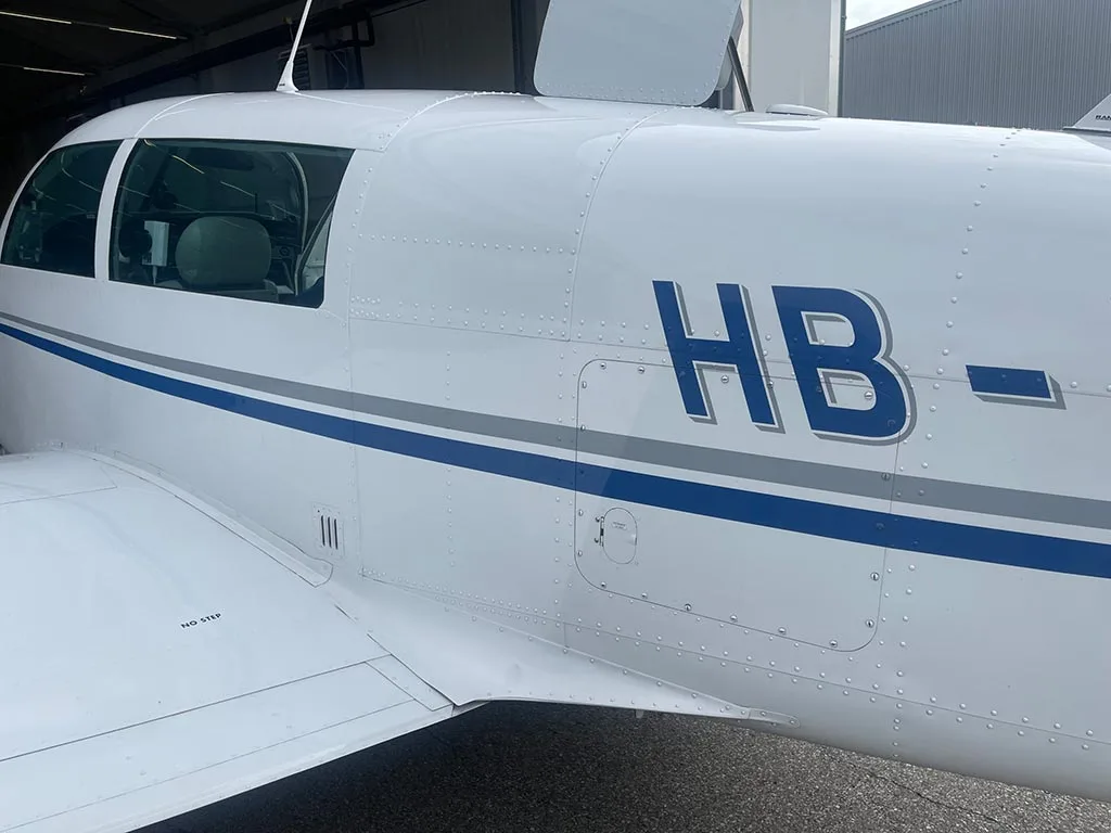 Close-up of the fuselage of a white aircraft with blue and grey stripes, showing part of the registration 'HB-'.
