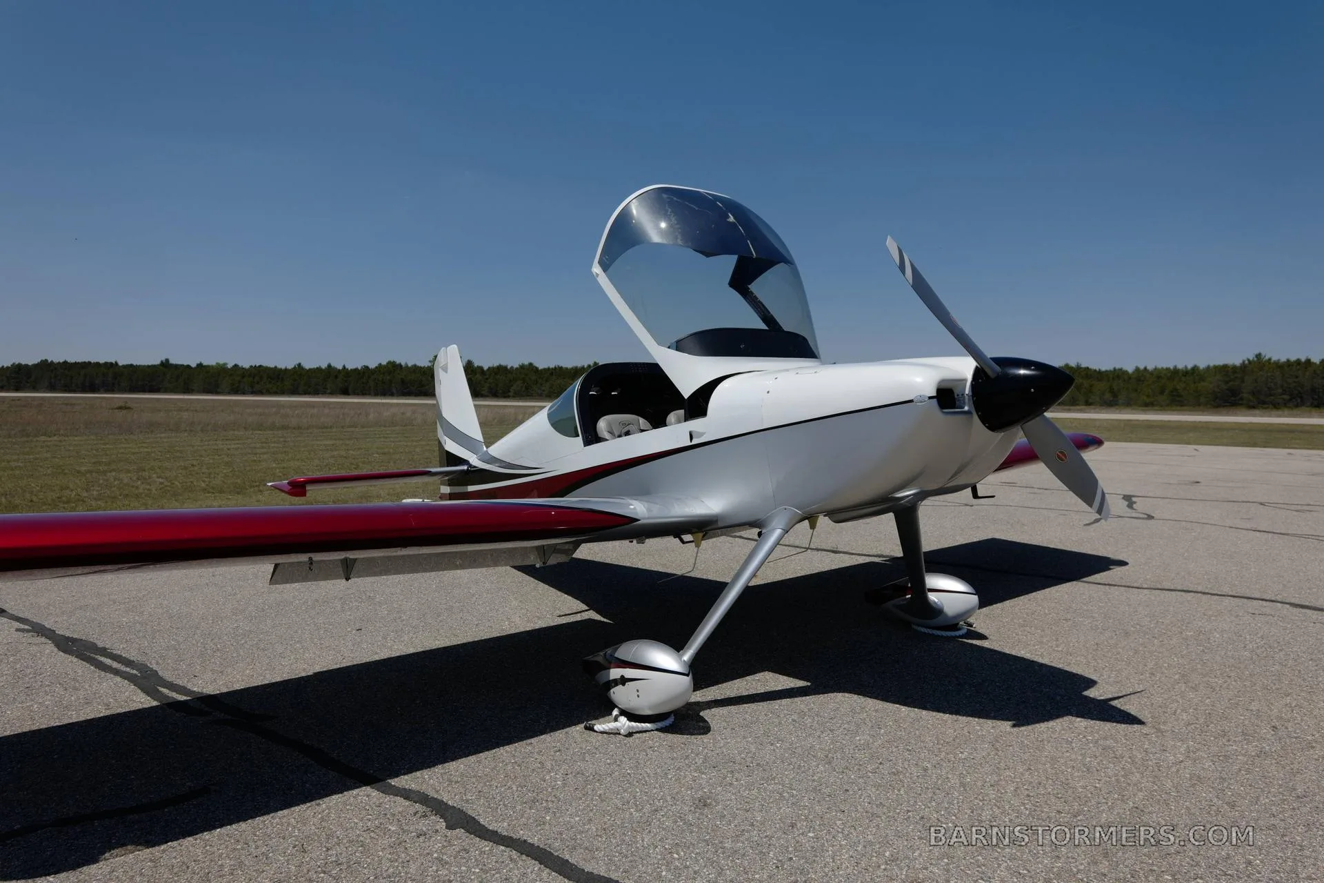 A white and red Vans RV-140MP aircraft with an open canopy, parked on an asphalt runway with a distant tree line.