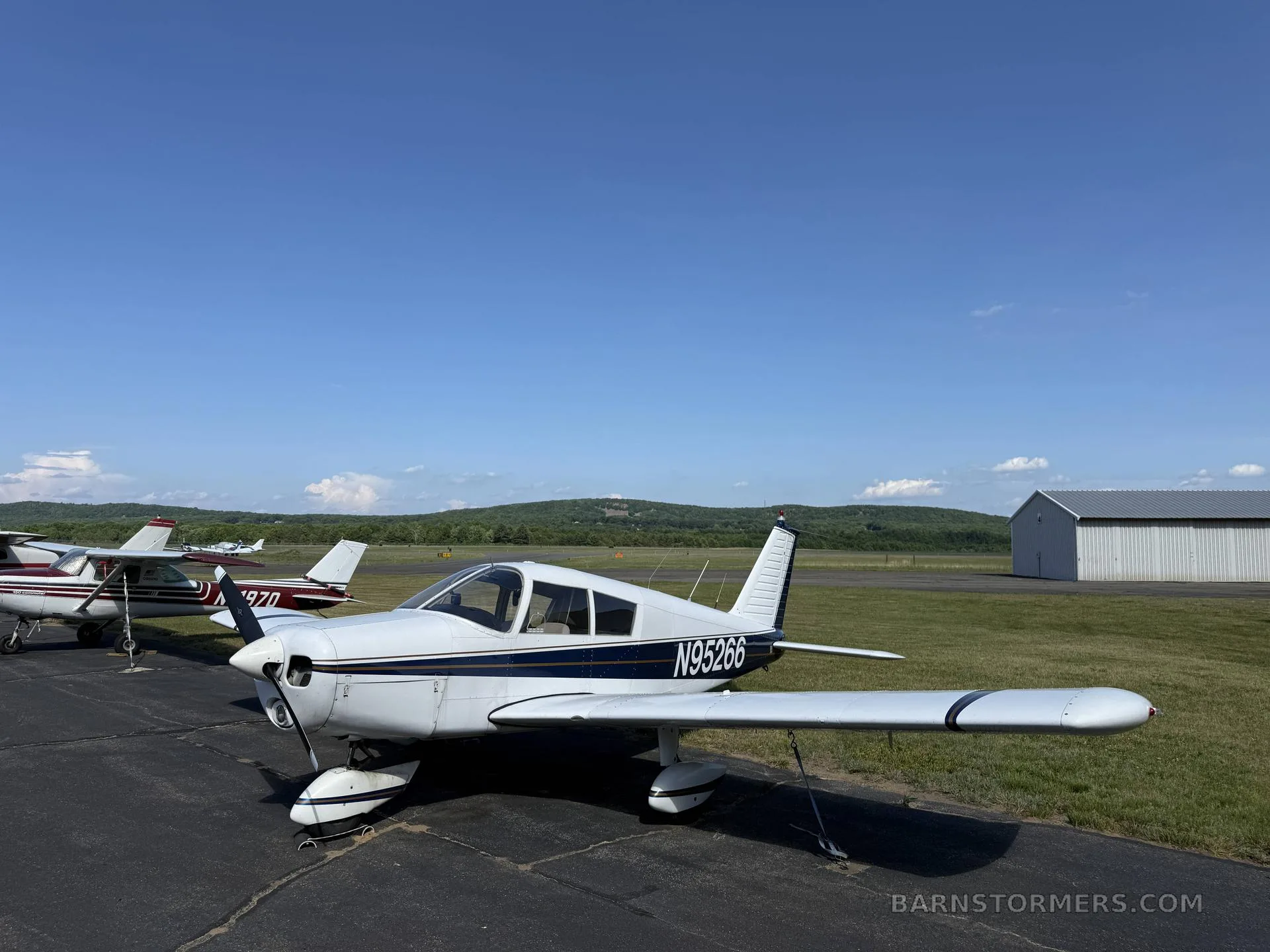 A white single-engine propeller aircraft (N95266) with blue stripes parked on an airport apron with other planes and a hangar.