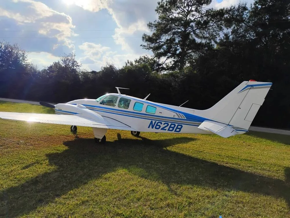 A white Beechcraft Baron N62BB with blue stripes is parked on a grassy field with trees in the background, viewed from the right side.