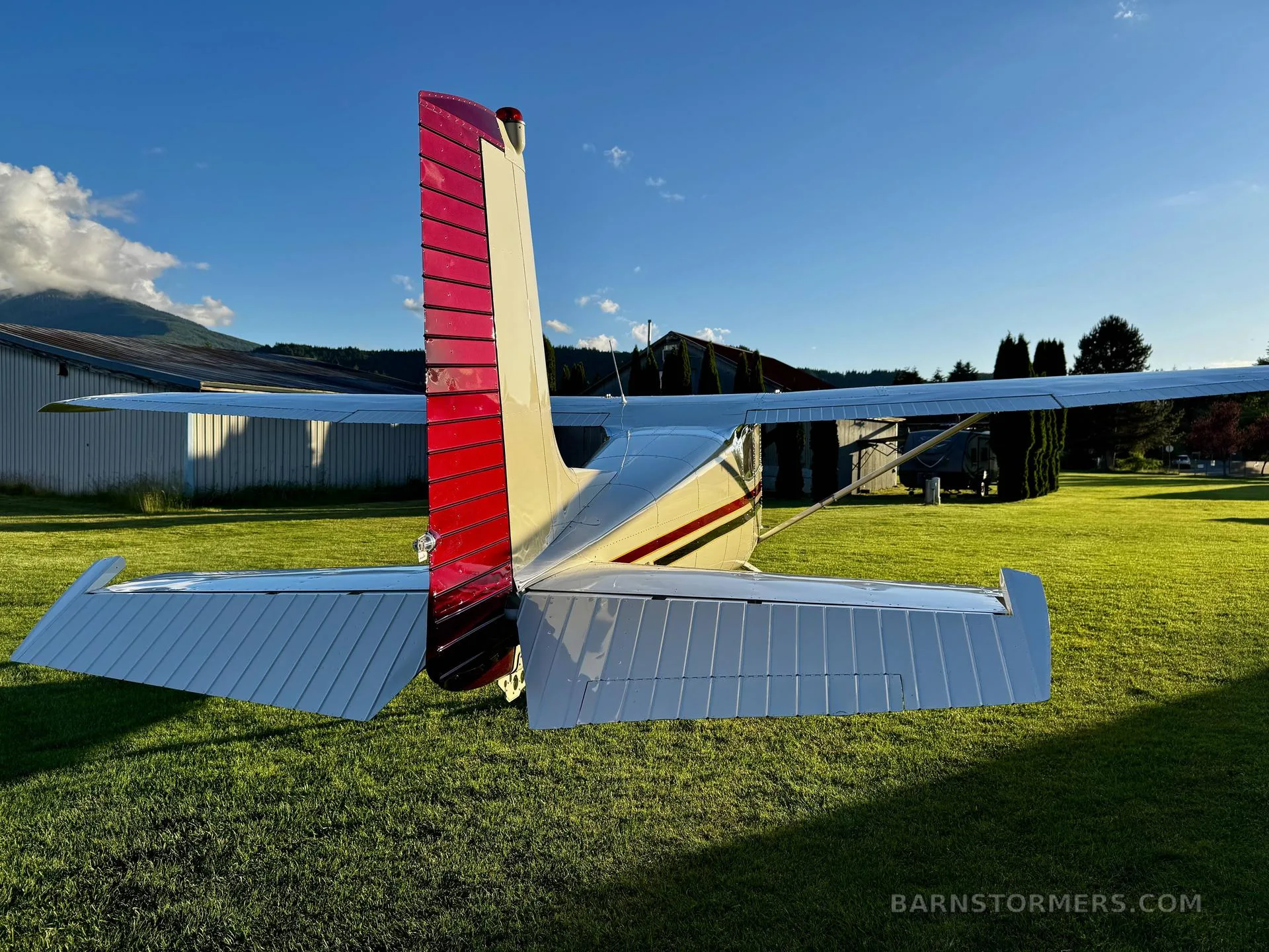 A rear view of a white aircraft with a red tail and white wings, parked on a green grassy field with hangars in the background.
