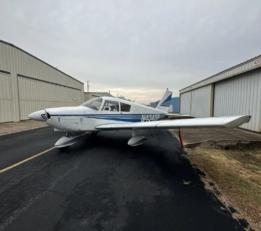 A white single-engine propeller aircraft with blue stripes (N4345P) is parked on asphalt between two hangars under an overcast sky.