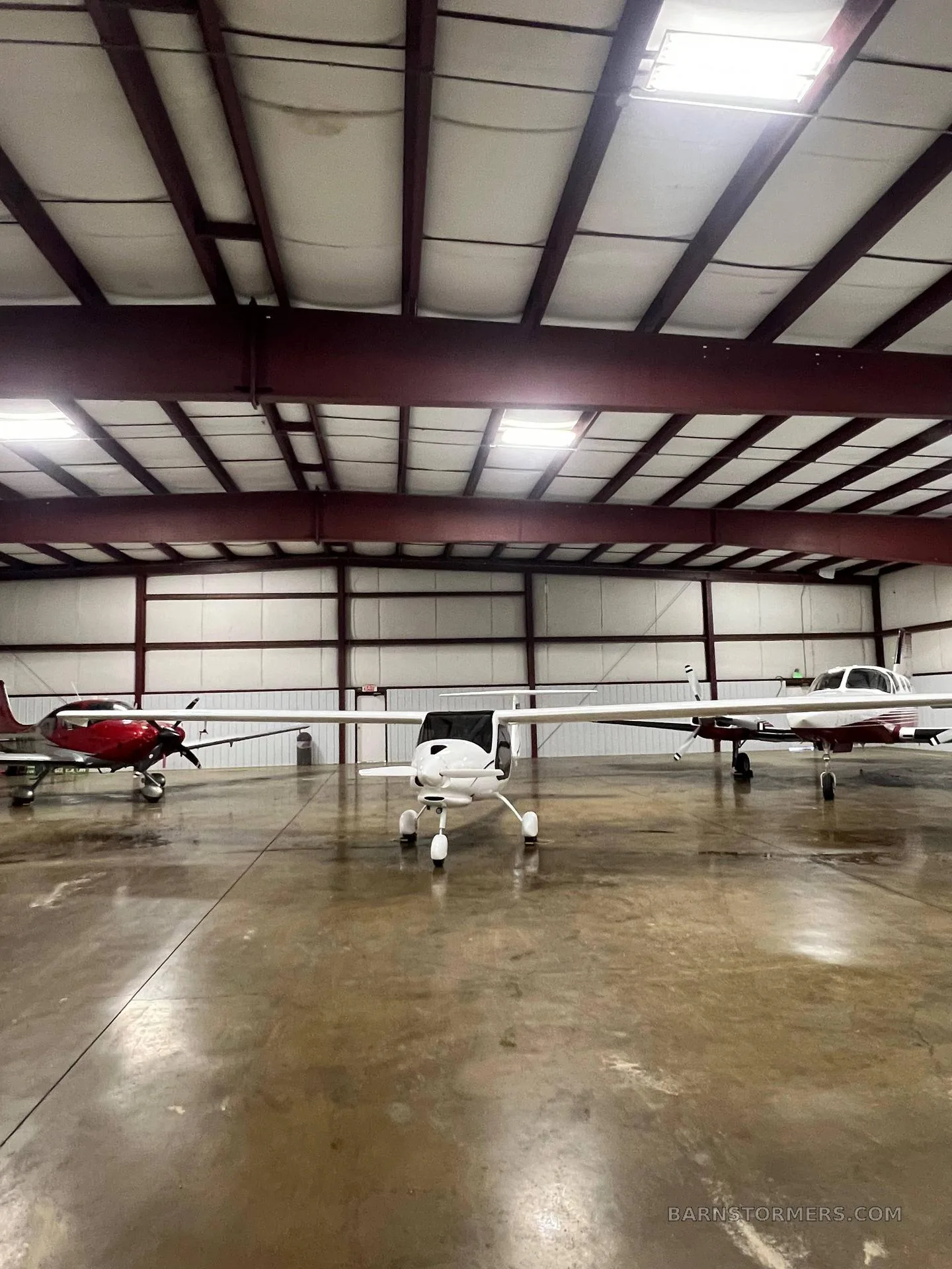 A white Pipistrel Sinus light sport aircraft is parked in the center of a hangar, flanked by a red single-engine aircraft and a white and maroon aircraft.