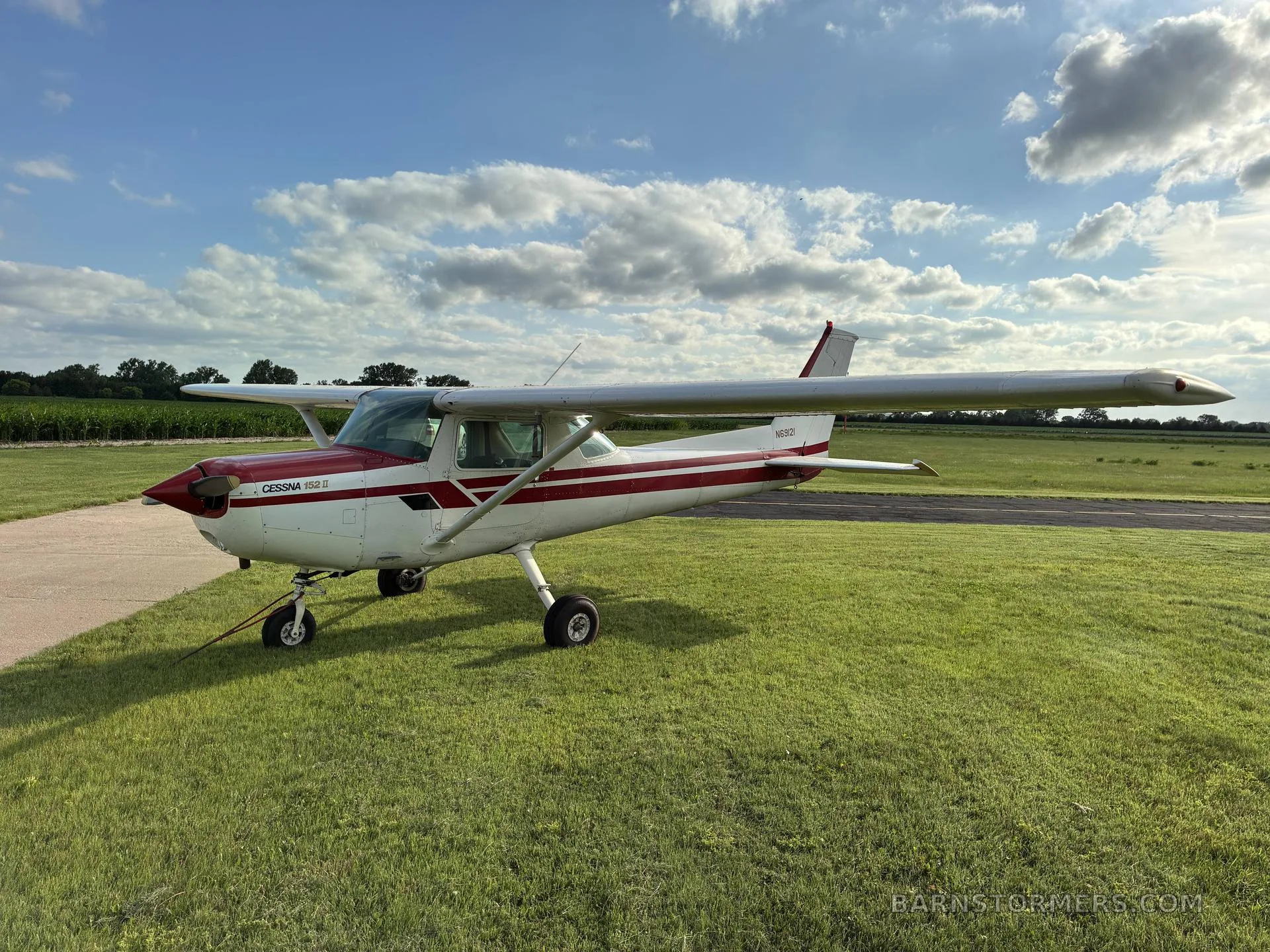 A white and red Cessna 172 aircraft is parked on a grassy field next to a runway under a partly cloudy sky.