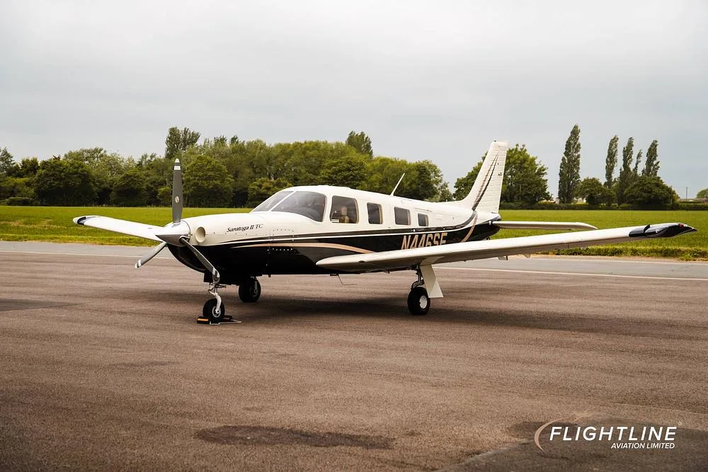 A white and black Piper Saratoga II TC, registration N446SE, parked on an asphalt runway with green fields and trees in the background.