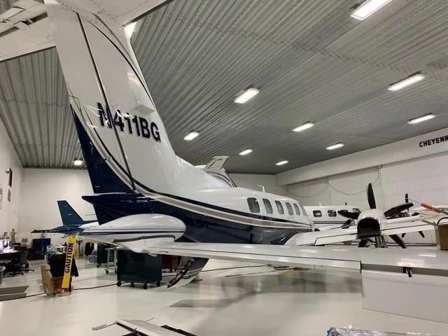 A white and blue twin-engine turboprop aircraft, likely a Beechcraft King Air, is parked inside a hangar, viewed from the rear-right.