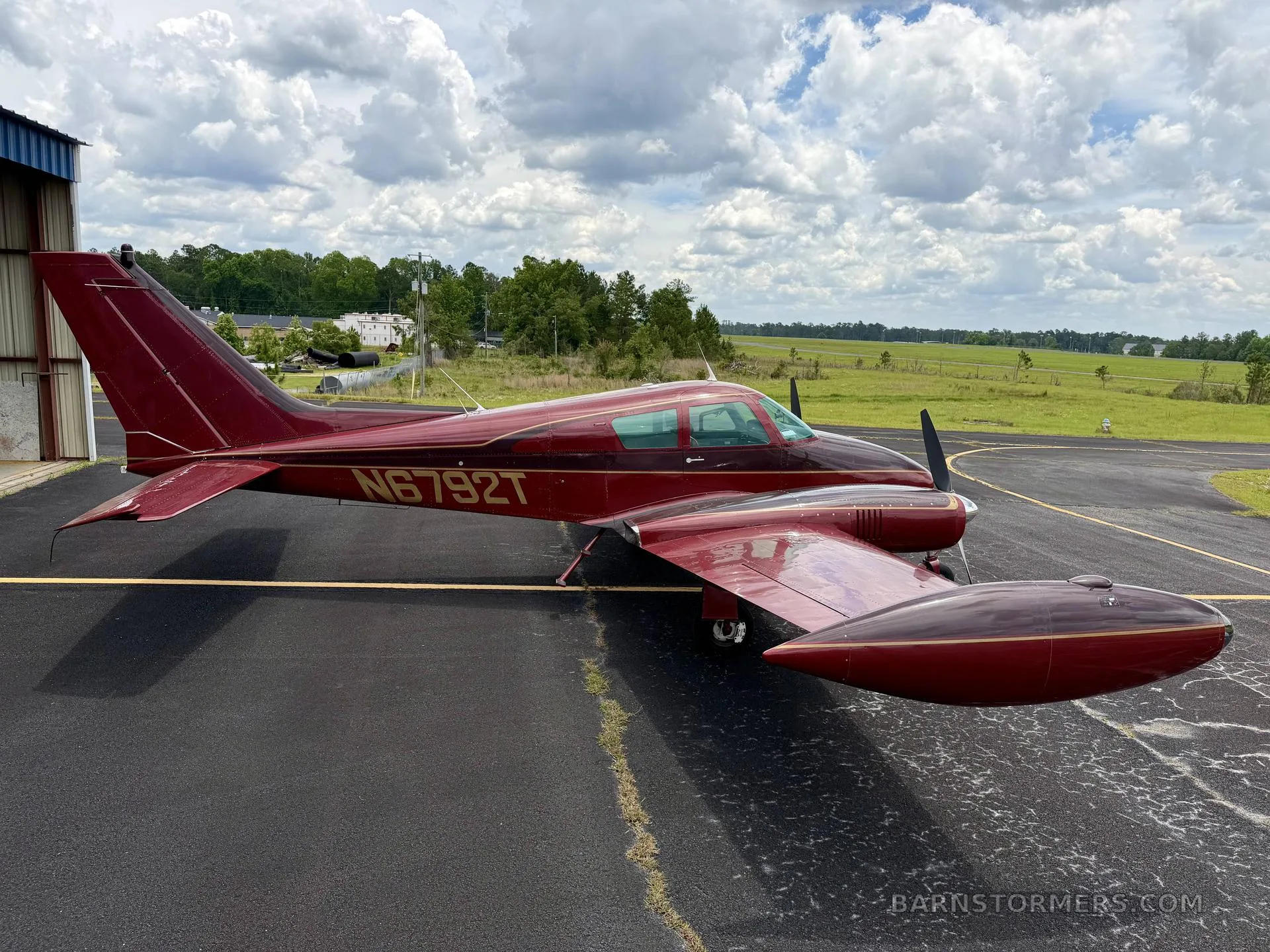 A red twin-engine aircraft, likely a Beechcraft Baron, with registration N6792T, parked on an asphalt apron with a grassy field in the background.