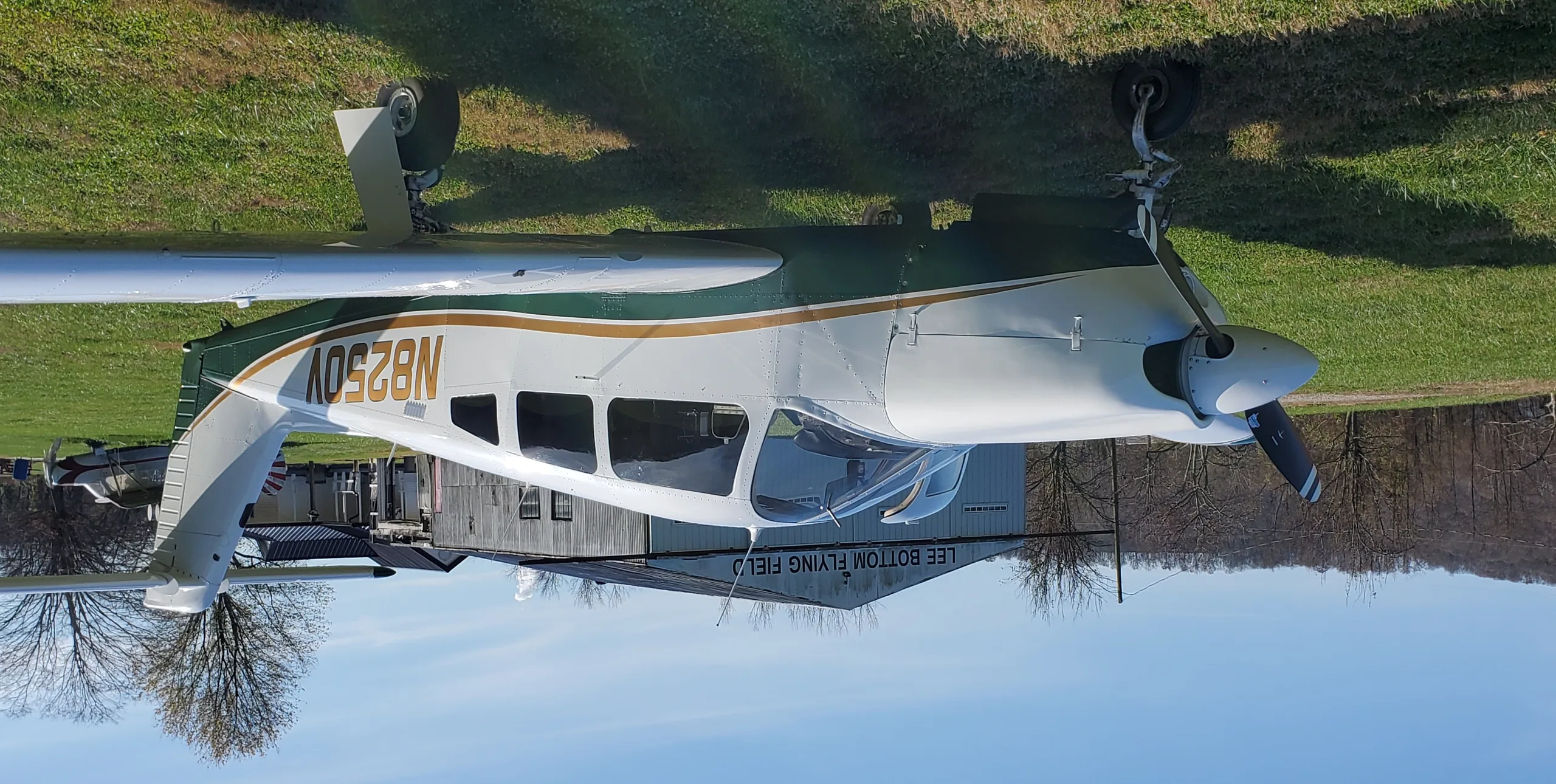 A white and green single-engine aircraft, N8250V, parked on a grassy airfield next to a building.