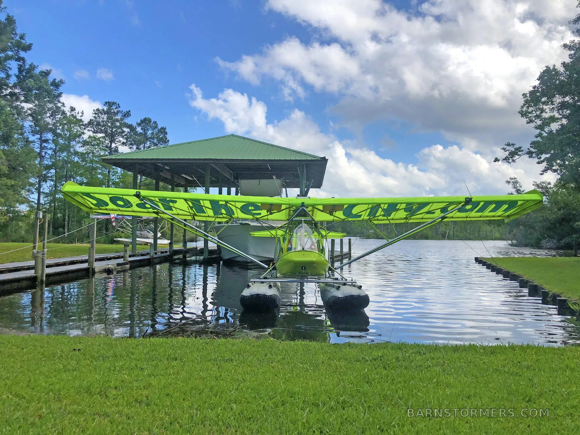 A bright green ultralight seaplane with "Soar The City.com" on its wings is docked in calm water next to a boathouse under a partly cloudy sky.