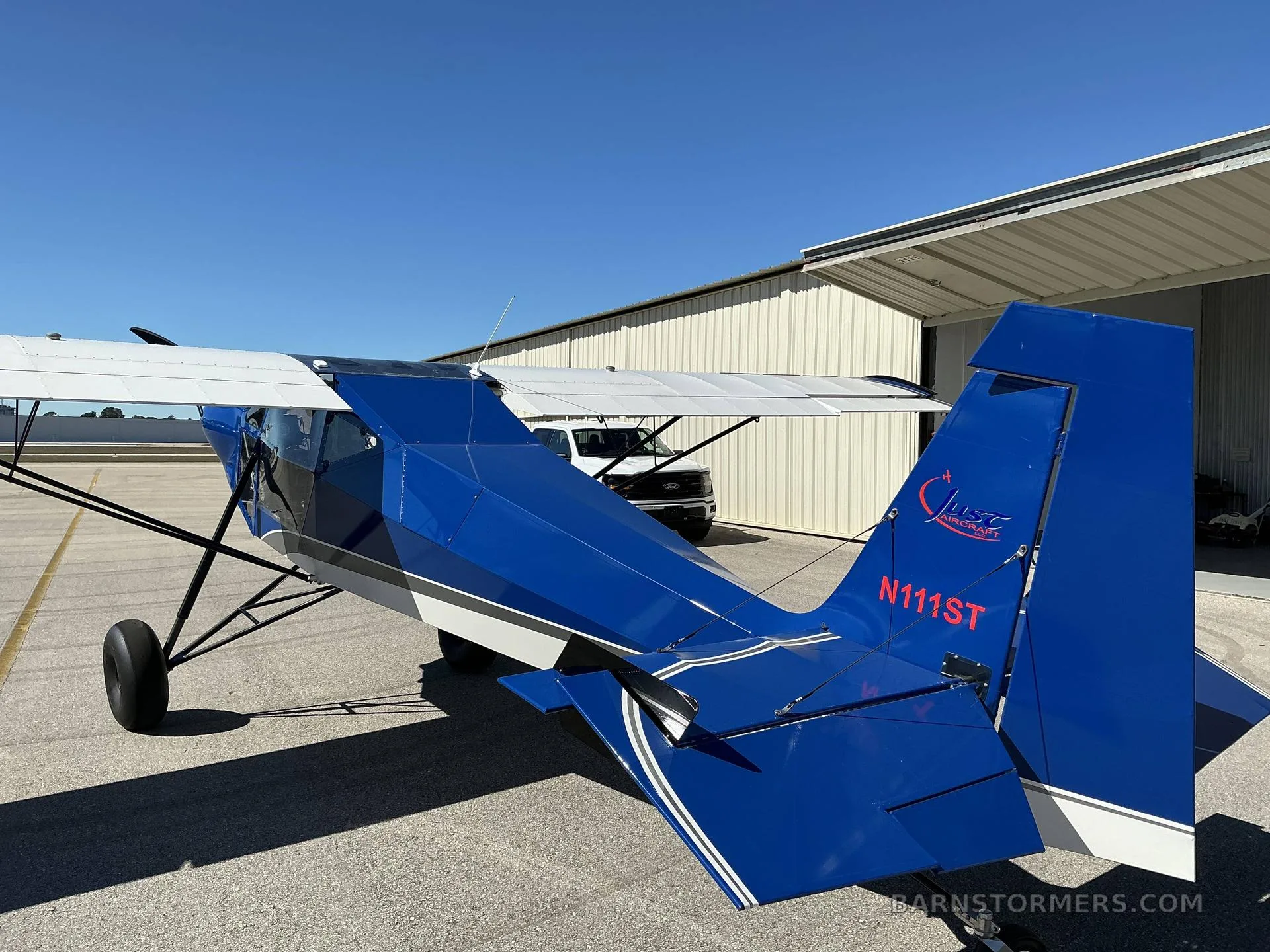 A rear-quarter view of the blue and white SuperSTOL aircraft showing its tail with registration N111ST and "Just Aircraft" logo.