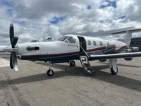 A white Pilatus PC-12 turboprop with dark red and gray stripes, parked on an airfield with its passenger door open.