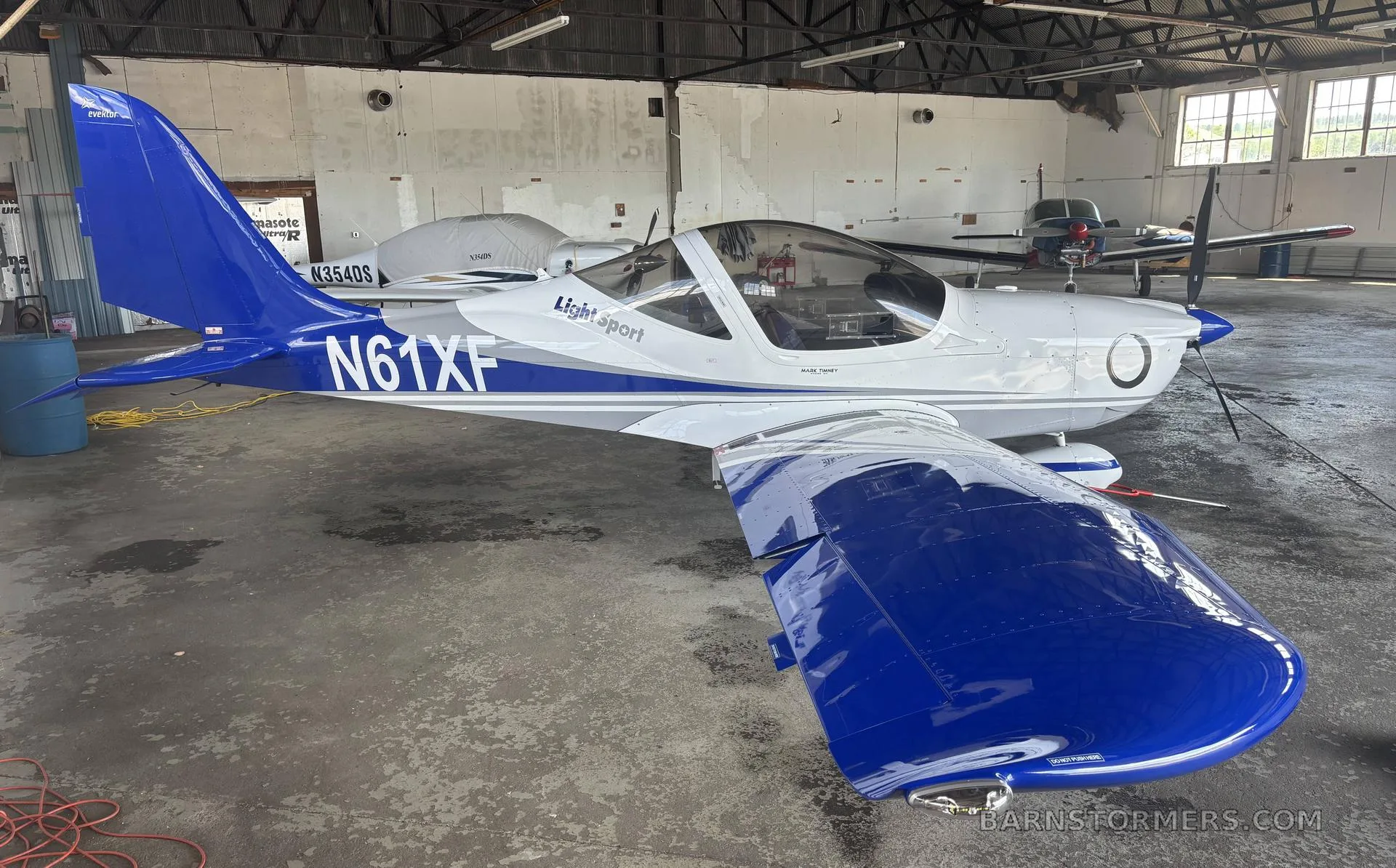 A white and blue Evektor Harmony Light Sport aircraft (N61XF) is parked inside a hangar.