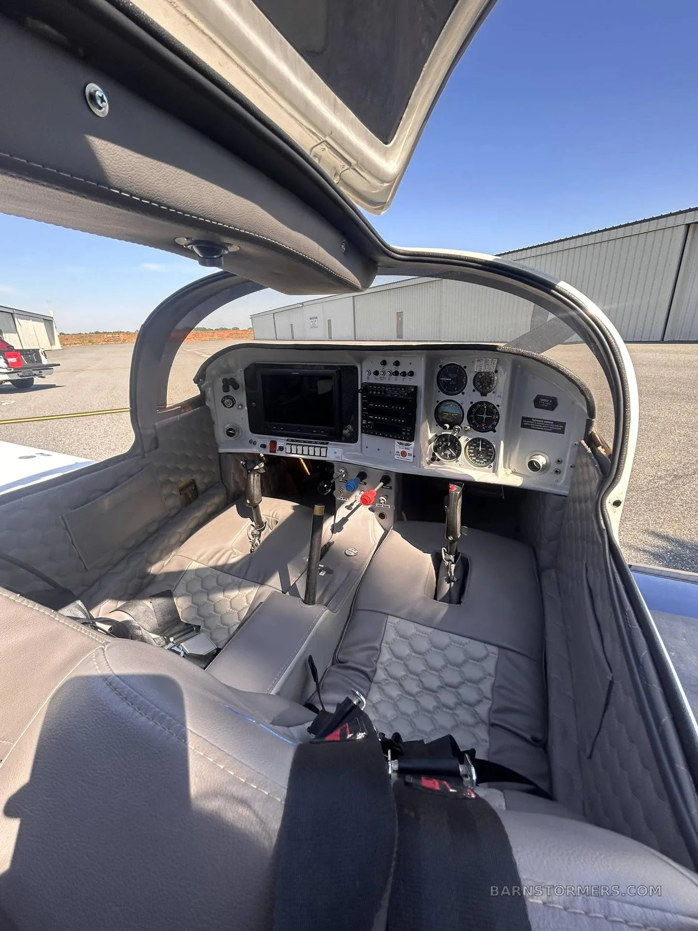 The cockpit of a small aircraft features a grey interior with a modern instrument panel and two seats, viewed with the canopy open.