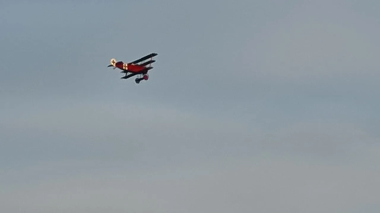 A red Fokker Dr.I replica triplane with German crosses on its wings, flying against a cloudy sky.