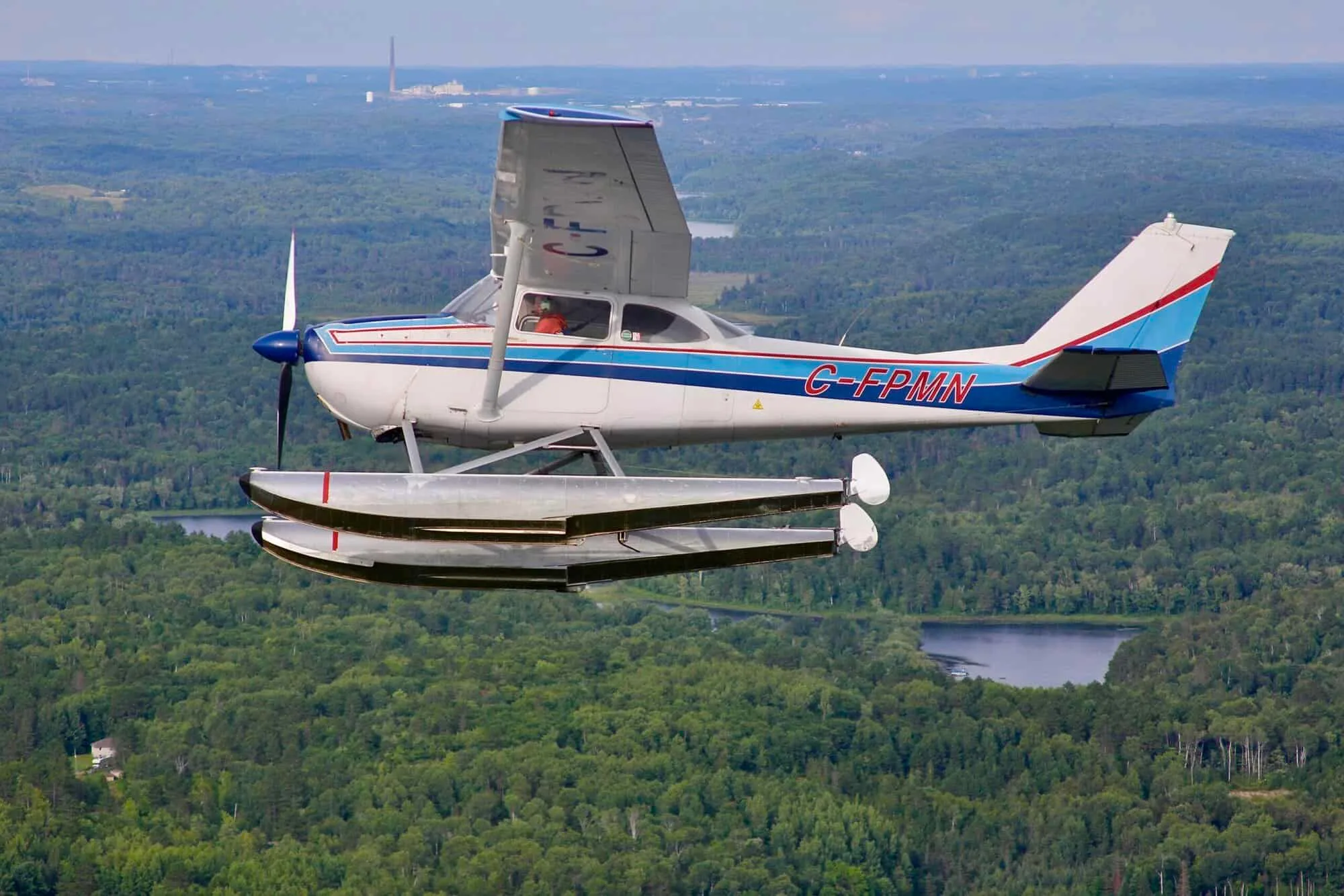 A white and blue Cessna 172 floatplane, registration C-FPMN, flies over a green forest and lakes.