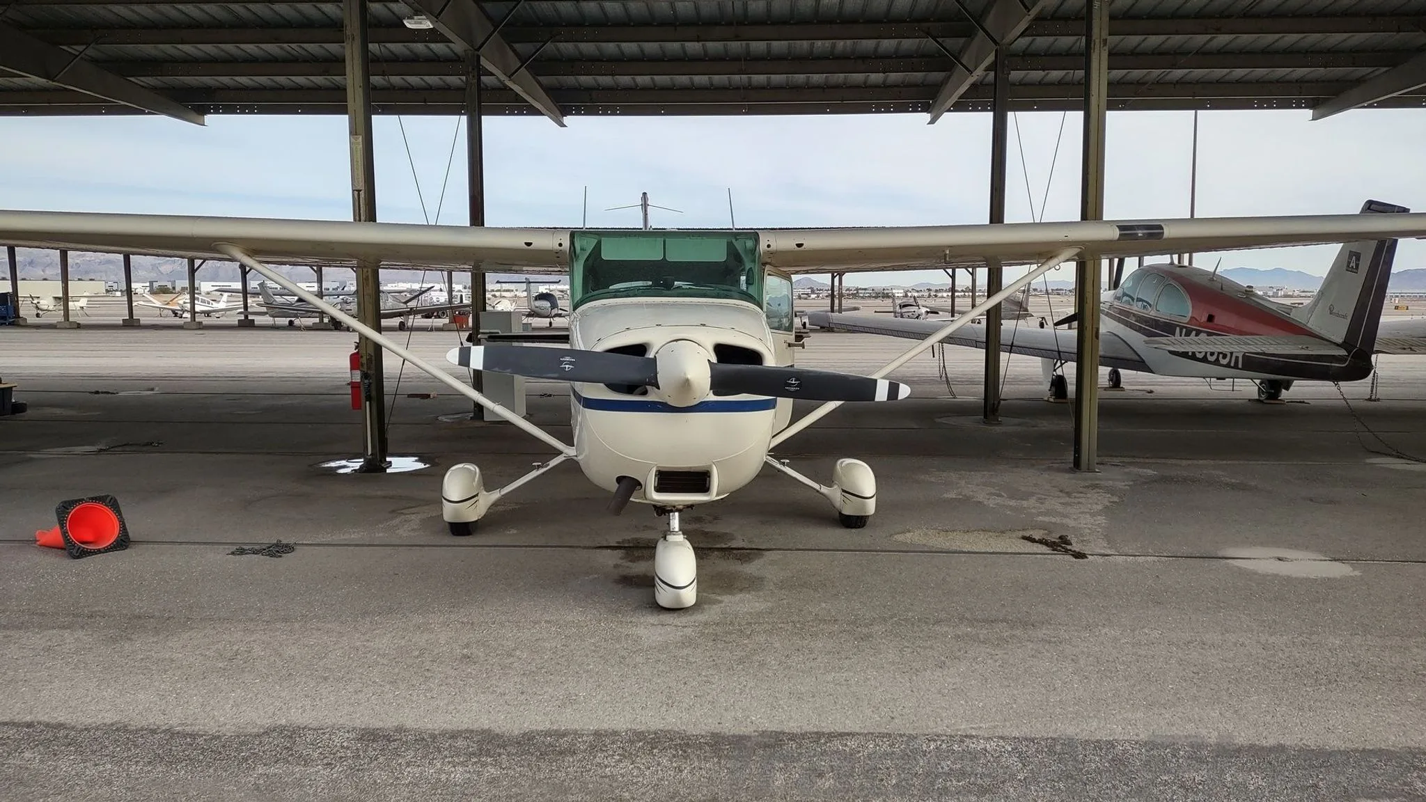 A white Cessna 172 with blue and red stripes parked head-on under a hangar, with other aircraft in the background.