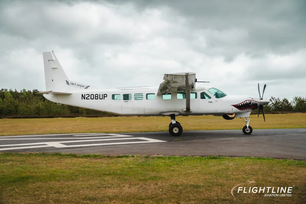 Side view of a white Cessna 208 Caravan with N208UP registration and shark teeth nose art, on a runway.