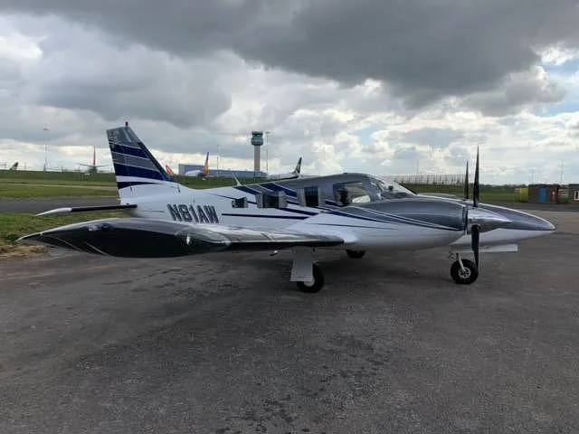 A white and grey twin-engine aircraft (N81AW) with blue stripes parked on an airfield with an air traffic control tower.