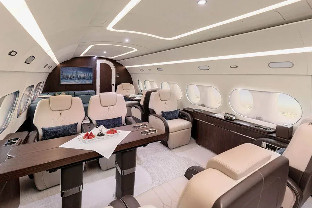A private jet cabin with light beige leather seats, a wooden table set with fruit, and a TV screen showing a snowy landscape.