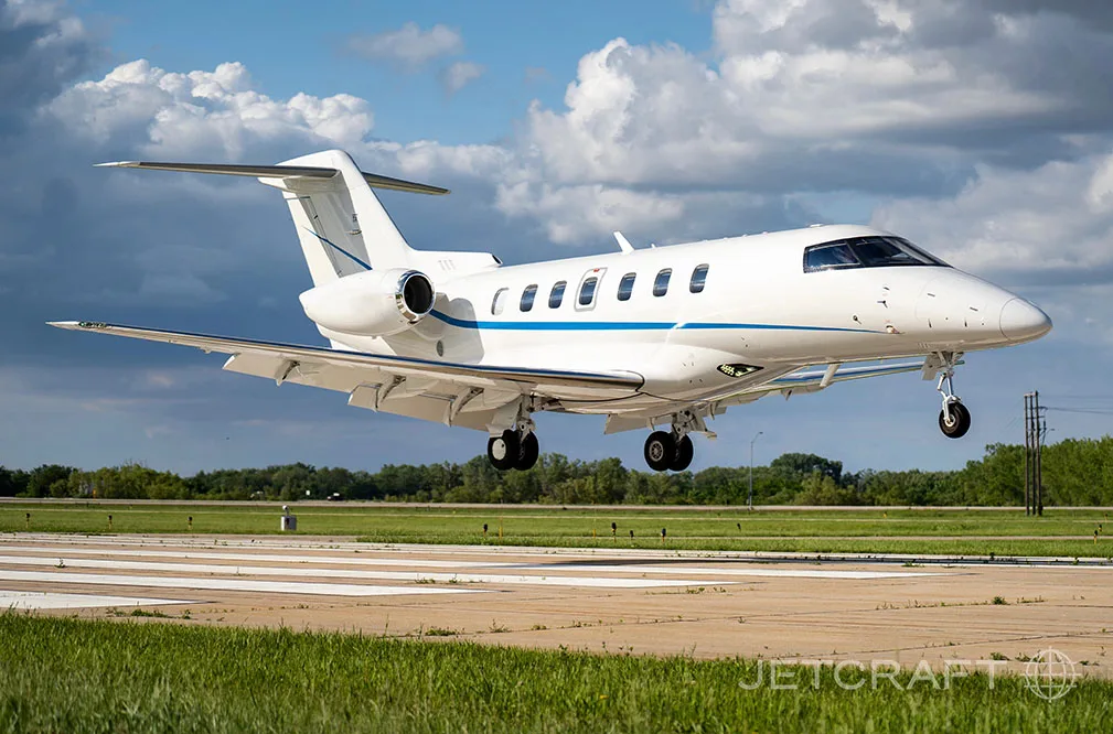 A white business jet with blue stripes is captured mid-air, just above a runway, with its landing gear extended.