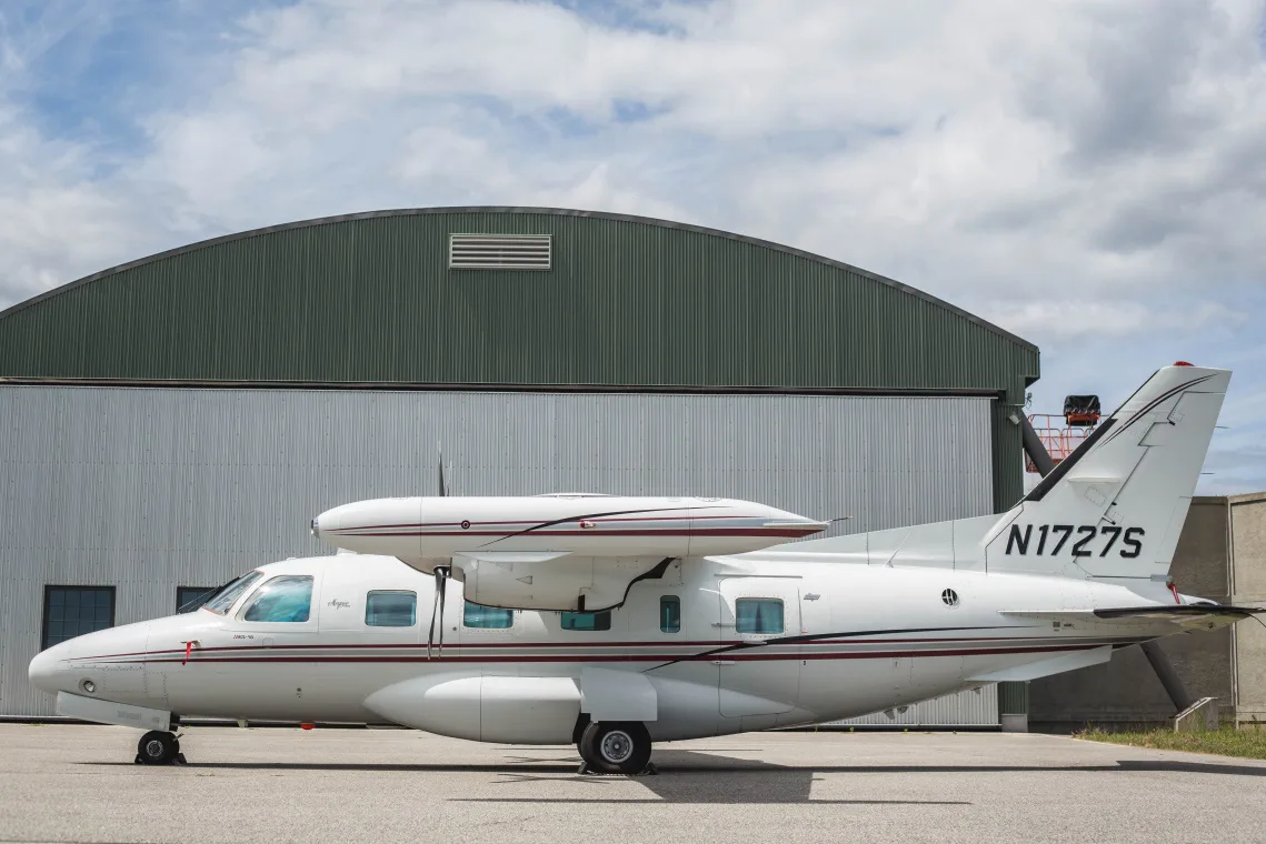 A white Piper Cheyenne aircraft with maroon stripes parked on an apron in front of a hangar under a cloudy sky.