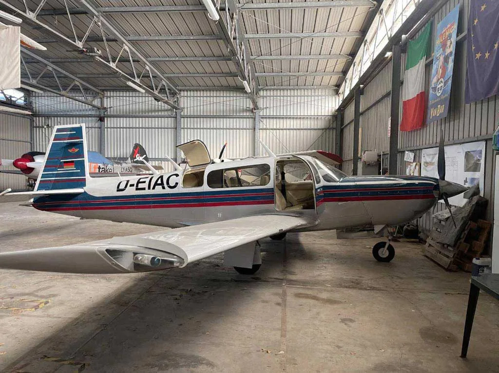 A Mooney M20J aircraft with its doors open, parked inside a hangar with flags hanging in the background.