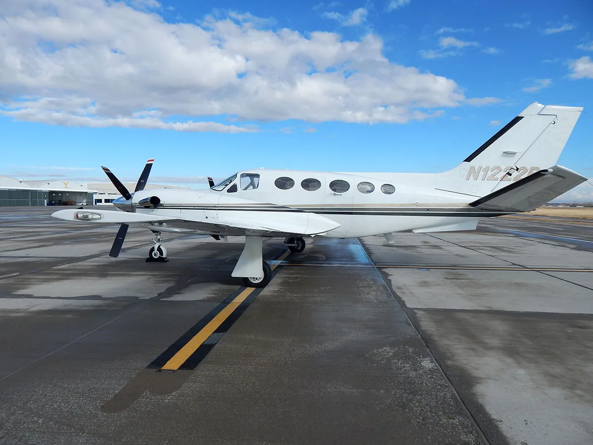 A white twin-engine turboprop aircraft, N1222P, with black and gold stripes, is parked on a wet tarmac under a partly cloudy sky.