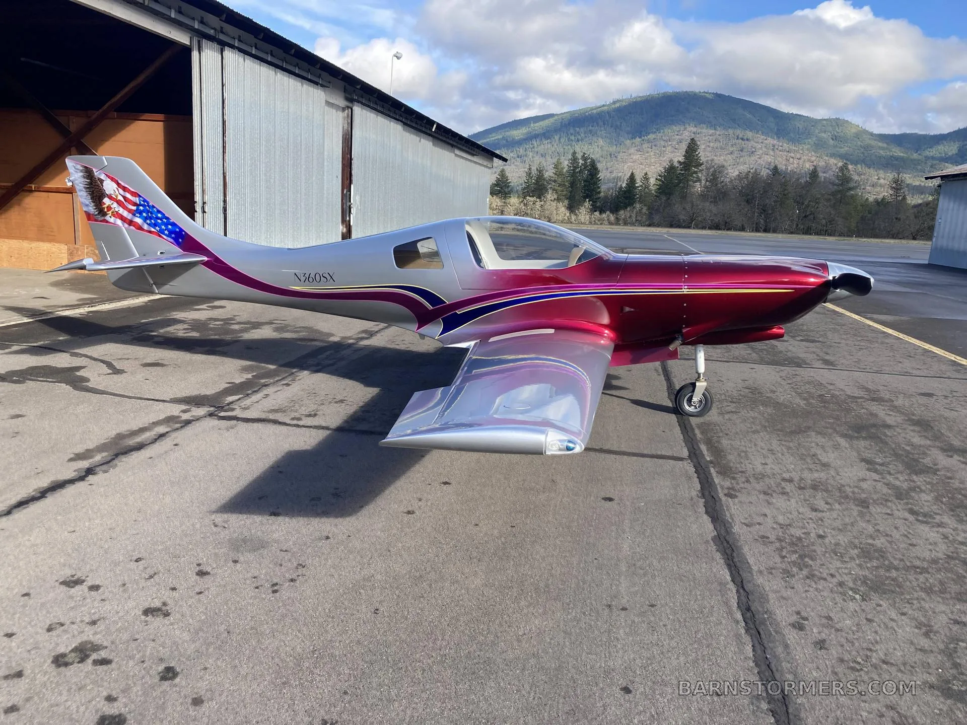 A silver and metallic red Lancair 360 aircraft with an American flag and eagle tail art, parked on an asphalt ramp in front of a hangar with mountains in the background.