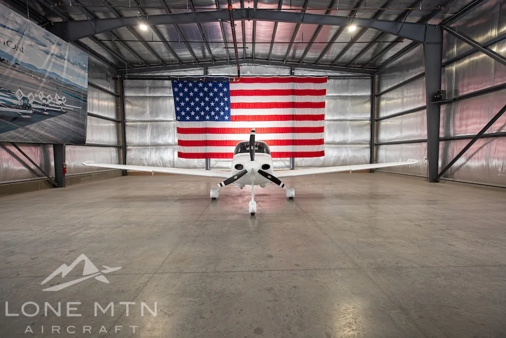 A white Cirrus SR22 aircraft with blue and black stripes, tail number N643LV, parked head-on in a hangar with an American flag in the background.