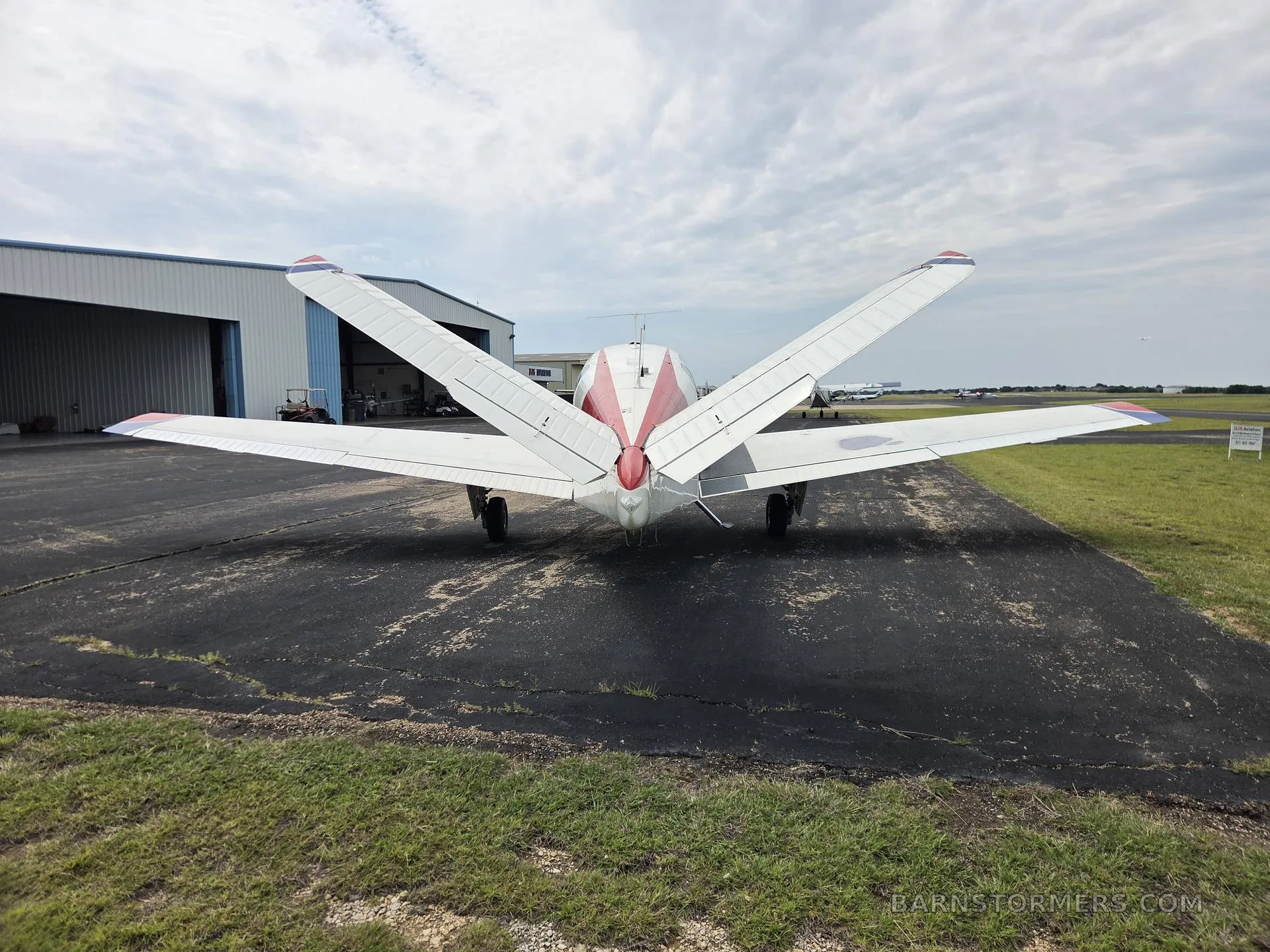 The distinctive V-tail of a white and red Beechcraft Bonanza aircraft is viewed from directly behind, parked on an asphalt ramp in front of a hangar.