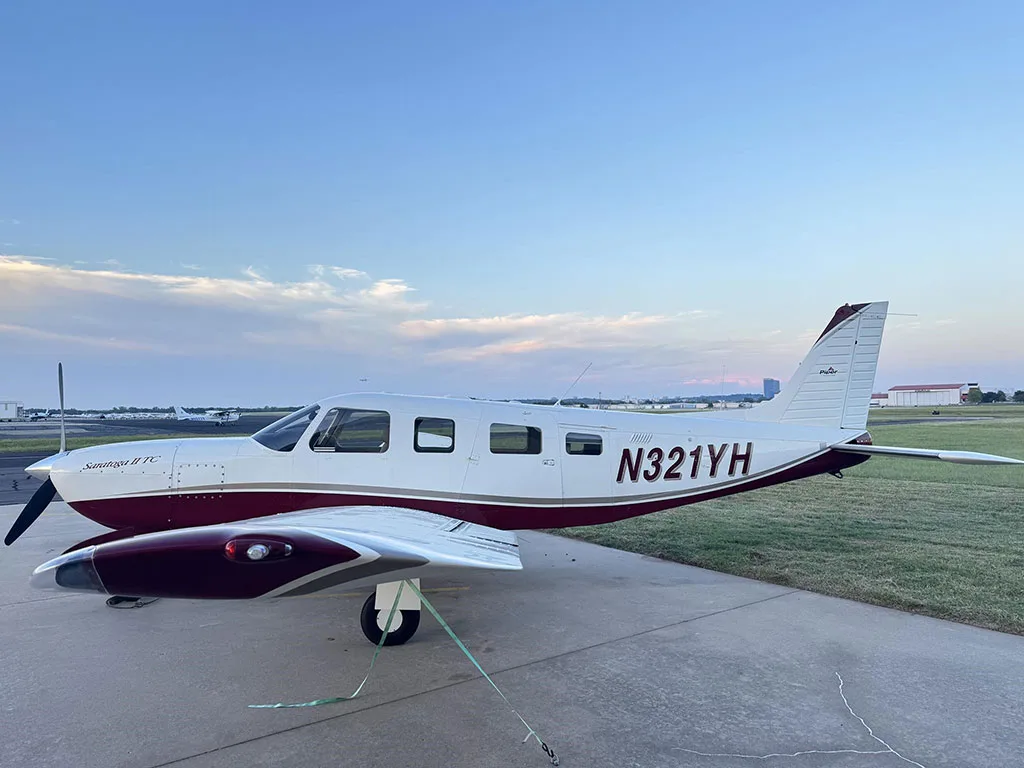 A white and maroon Piper Saratoga II TC aircraft (N321YH) tied down on an airport apron from the front-right view.
