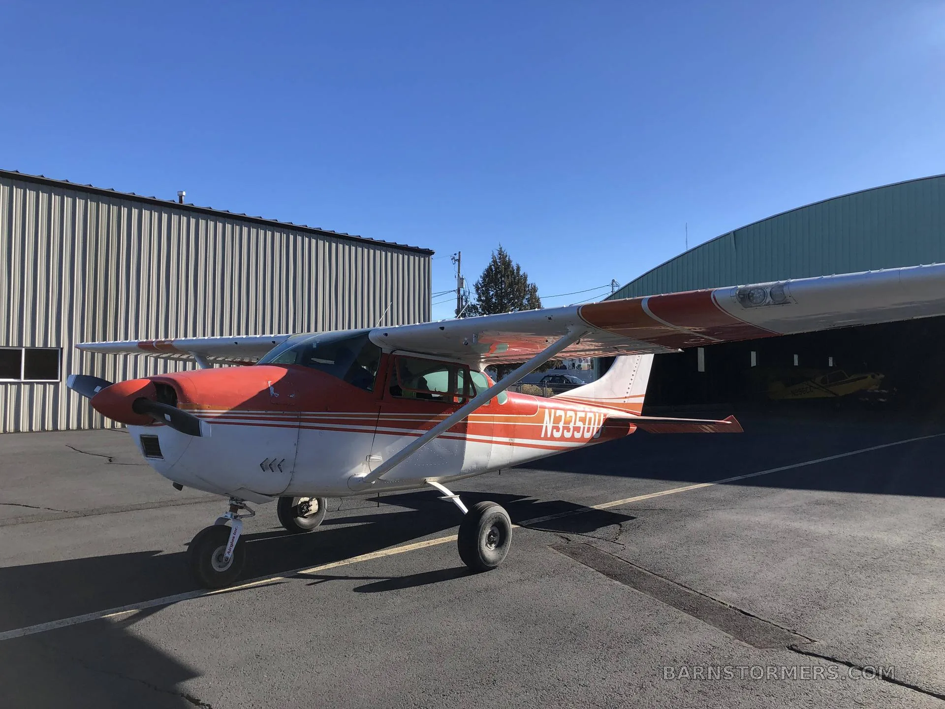 A red and white Cessna 182 Skylane (N3350U) is parked on an asphalt apron outside a metal hangar.