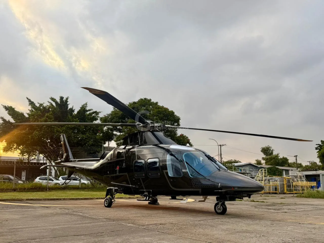 A black AgustaWestland AW109 helicopter parked on an apron under a cloudy sky.