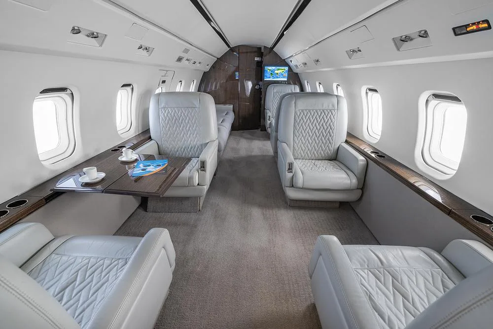 The elegant interior cabin of a business jet shows light gray leather seats, a table with coffee cups, and a view towards the front.