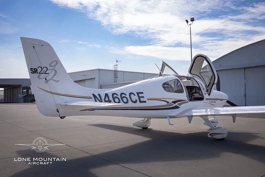 A white Cirrus SR22 aircraft with gold and black stripes, registration N466CE, parked on an airfield with its doors open, viewed from the rear-left.