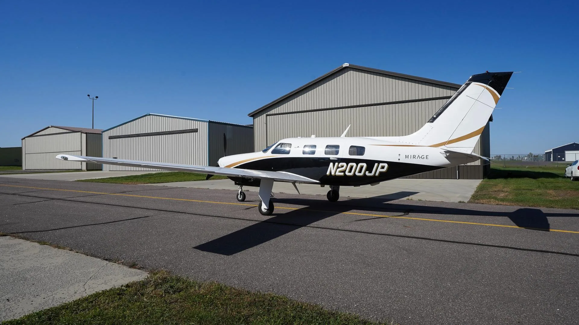 A white and black Piper Mirage N200JP with gold accents parked on an airfield next to hangars.