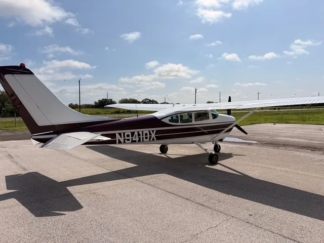A rear-left view of a white and maroon Cessna 182 Skylane (N9410X) on an airfield under a partly cloudy sky.