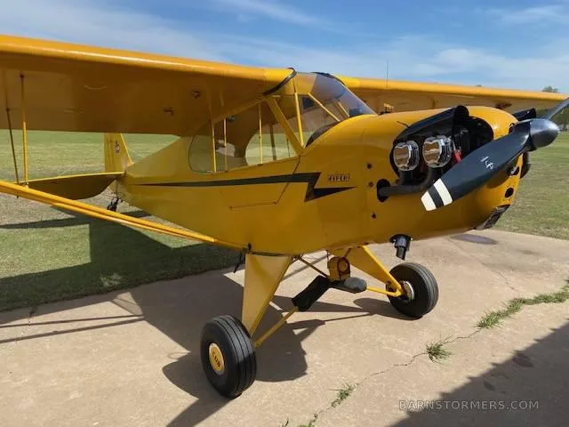 A three-quarter front view of a yellow Piper Cub-style aircraft with a black propeller, parked on a concrete pad.