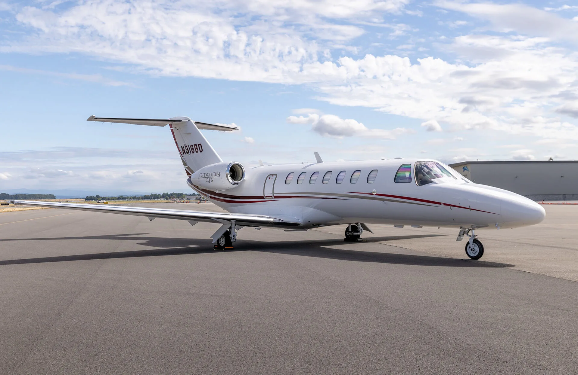 A white Cessna Citation CJ3 business jet with red and grey stripes is parked on an airport tarmac under a partly cloudy sky.