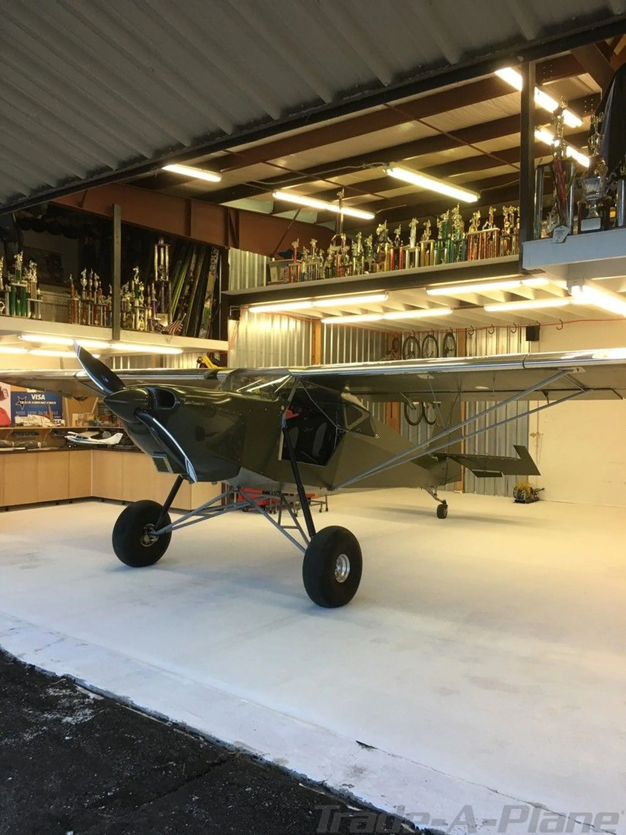 A green experimental aircraft with large tires sits inside a hangar filled with trophies.