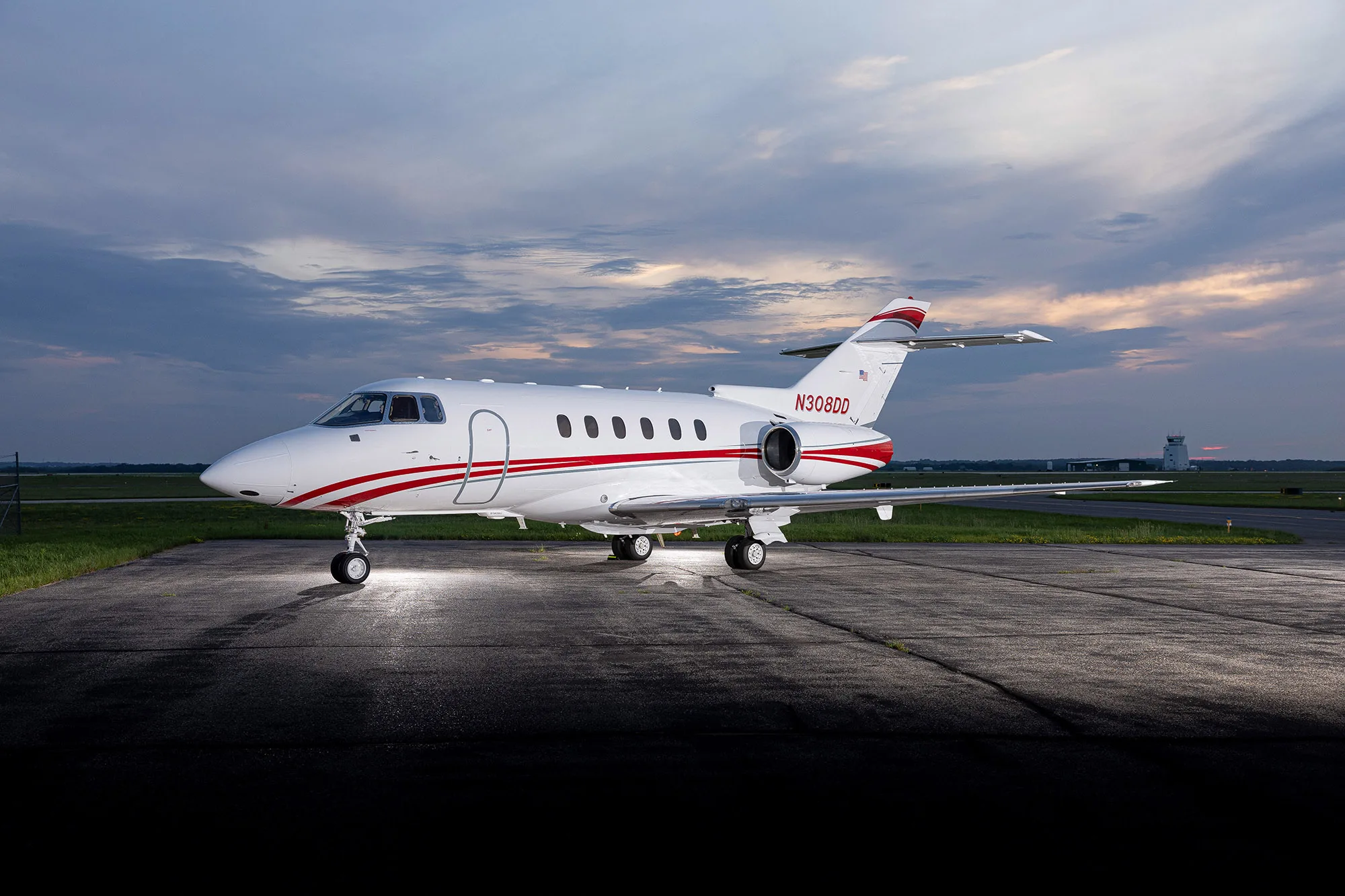 A white business jet with red stripes, registration N308DD, parked on an airfield at dusk, viewed from the front-left.