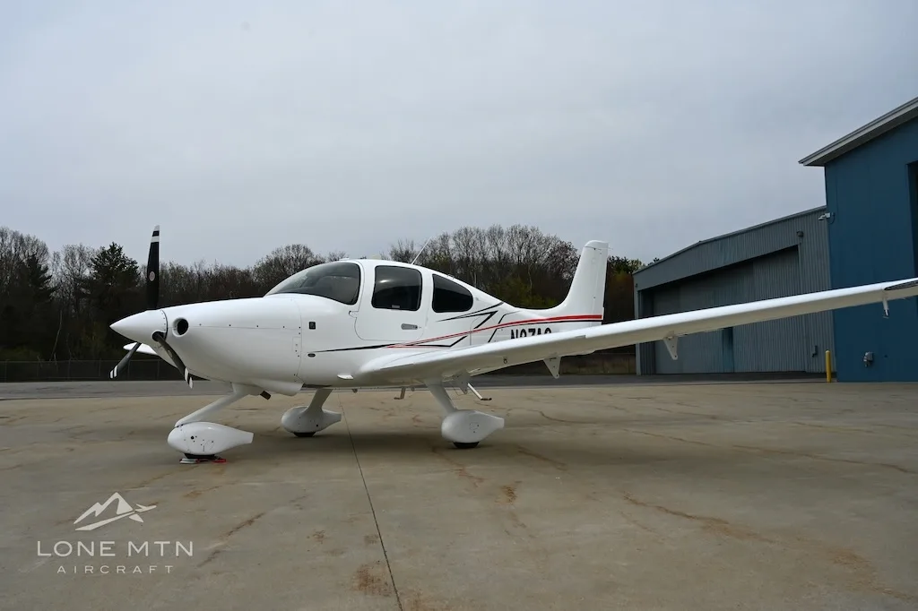 White Cirrus SR22 aircraft with red and black stripes, tail number N1071A, parked on an apron with trees in the background.