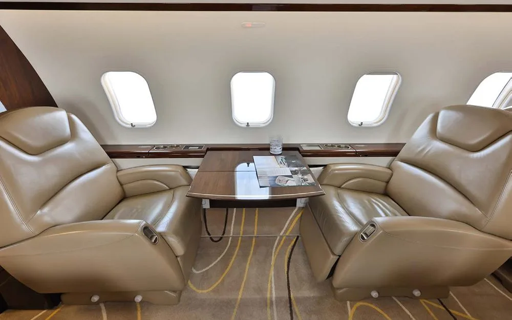 Two tan leather executive seats face each other with a polished wooden table in between, next to three aircraft windows.