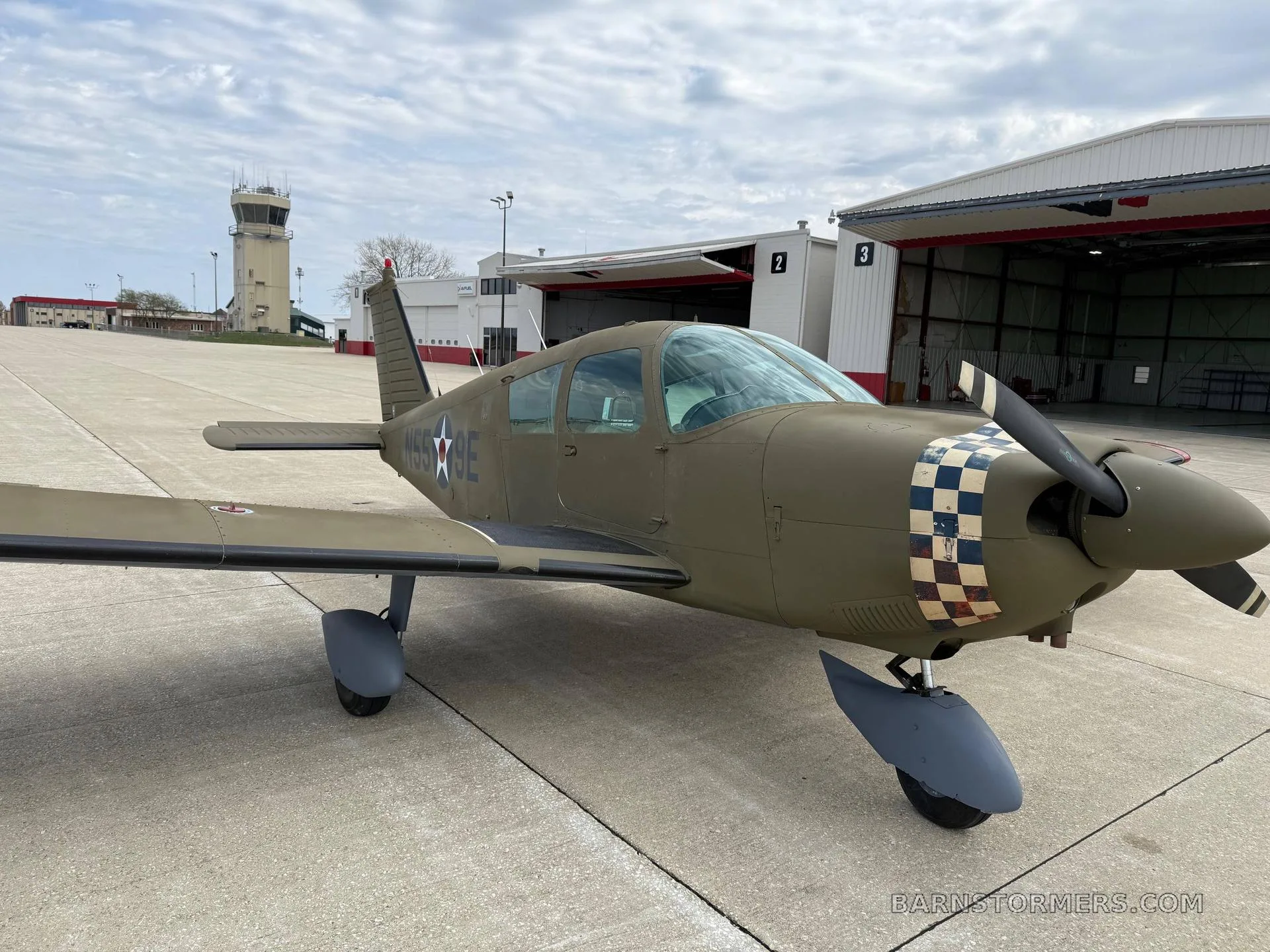 A green single-engine propeller aircraft with a checkered nose, parked on an airfield in front of hangars and a control tower.