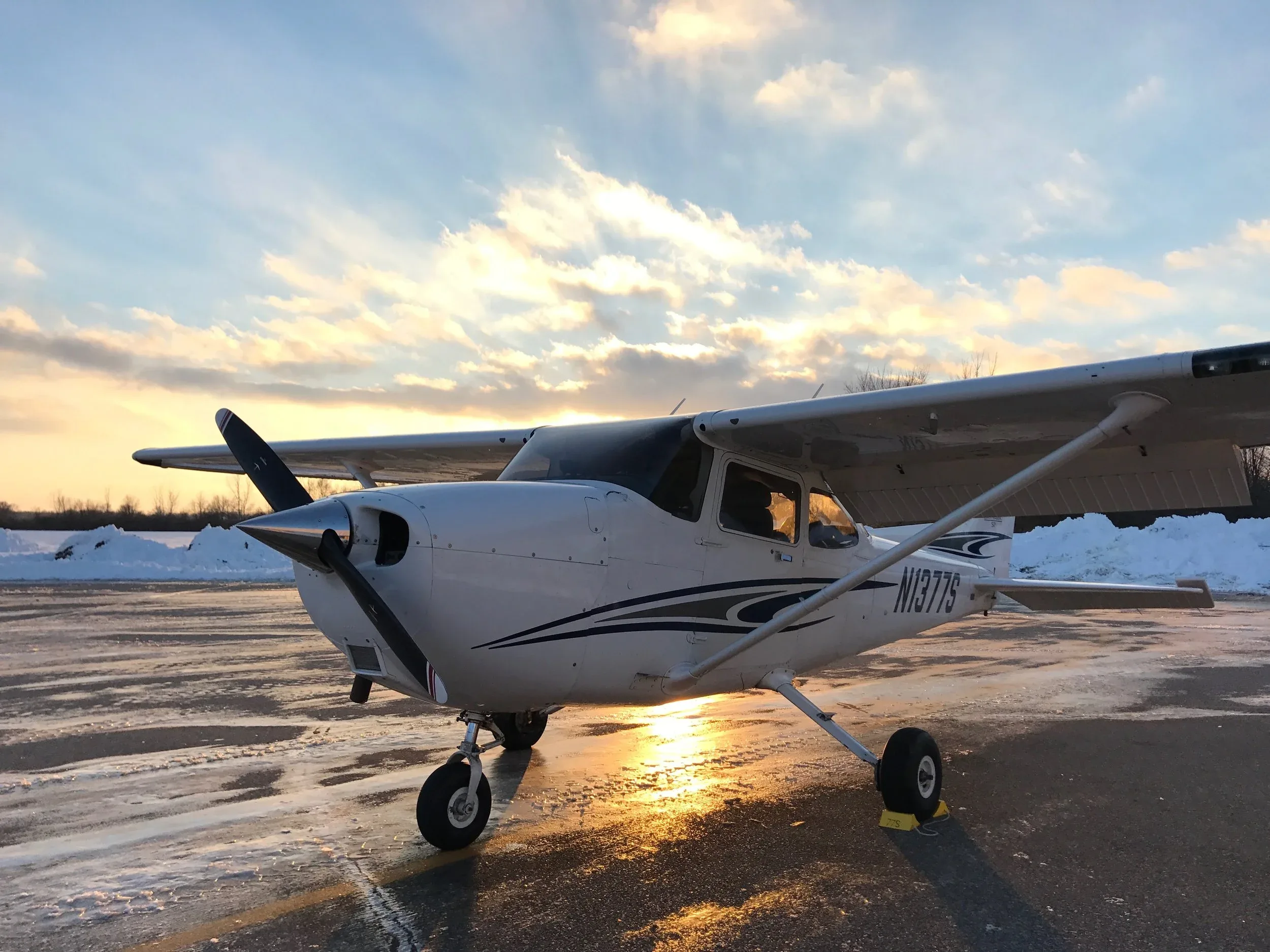 A white single-engine propeller aircraft with blue and black stripes on a wet tarmac at sunset, with snow in the background.