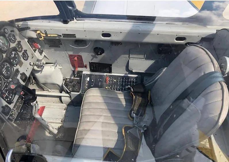 The interior cabin of an aircraft, showing two grey seats with harnesses and some side panel controls.