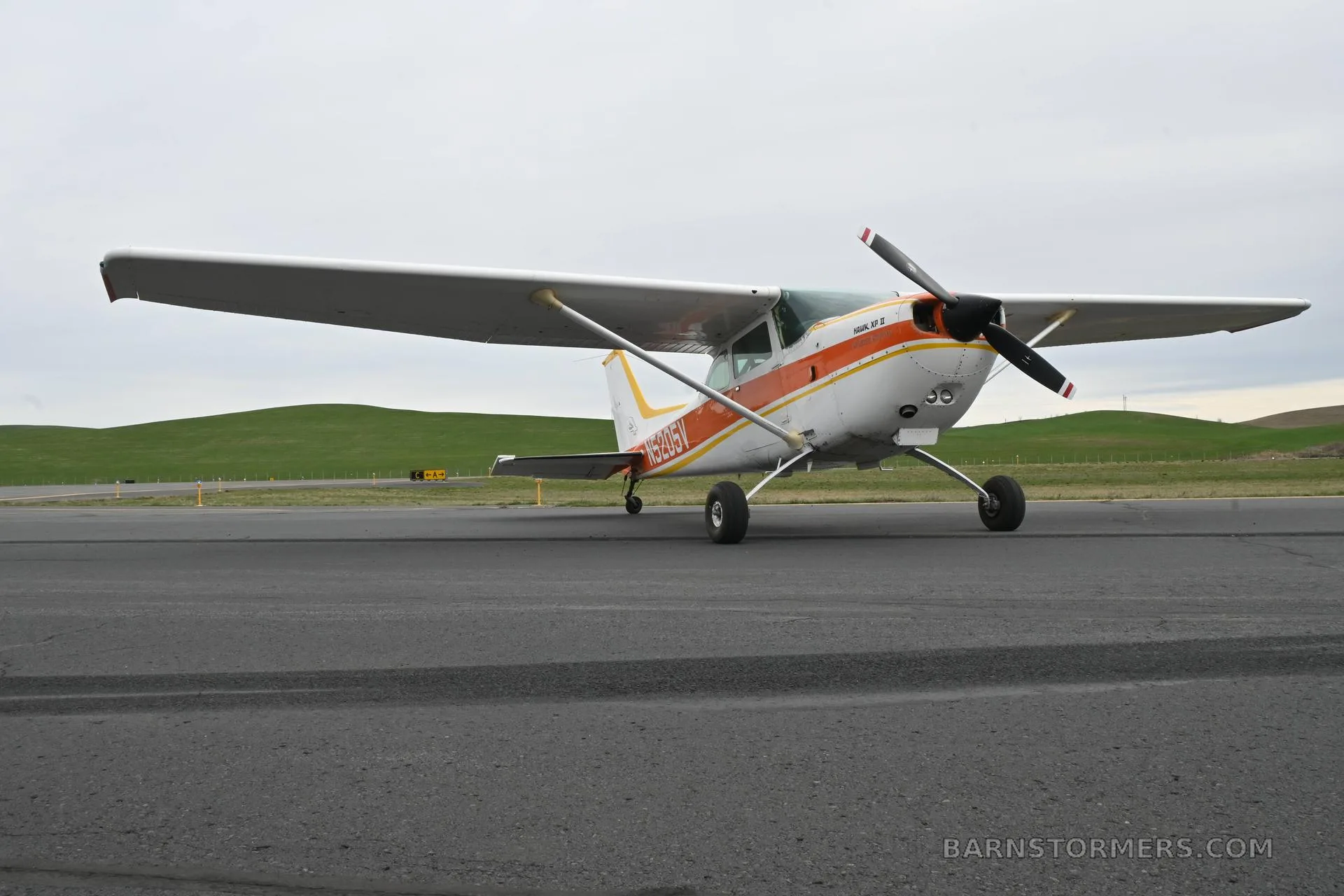 A low-angle front-side view of a white and orange Cessna 172 Hawk XP II (N5205V) on an asphalt runway.